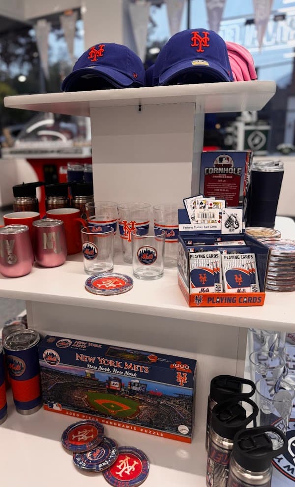 Tiered white retail display featuring New York Mets baseball caps, glassware, playing cards, and puzzles.